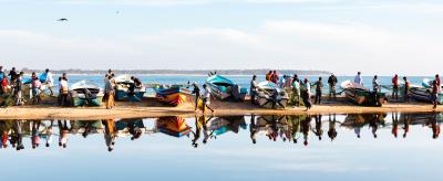 A group of people stands near several boats lined up on a sandy shore, with their reflections visible in the calm water. The sky is clear with a few clouds.