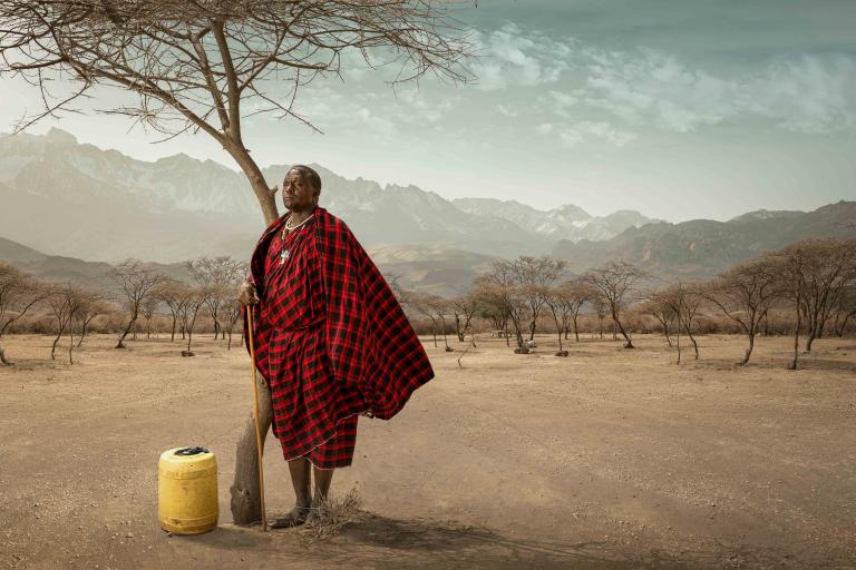 A person in traditional red attire leans against a tree in a dry landscape, with mountains in the background and a yellow container nearby.