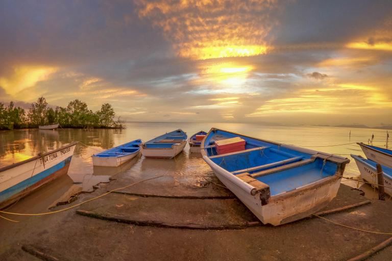 Three boats rest on a shore at sunset, with a vibrant orange and blue sky reflecting on the calm water, creating a tranquil and serene atmosphere.