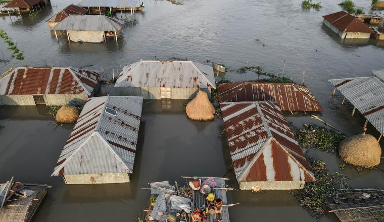 Aerial view of flooded village with people in a boat-like structure surrounded by partially submerged houses and muddy water.
