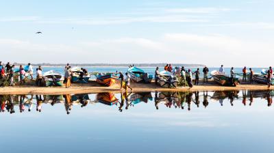 A group of people stands near several boats lined up on a sandy shore, with their reflections visible in the calm water. The sky is clear with a few clouds.