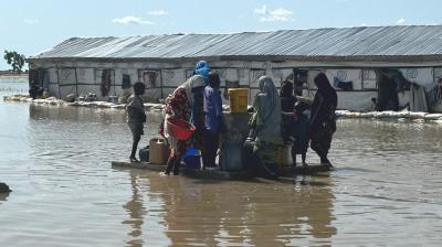 People gather with containers on a raft in floodwater near a building with a metal roof.