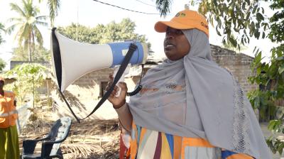 A woman in a gray headscarf and orange cap speaks into a megaphone outdoors, near a tree and some debris.