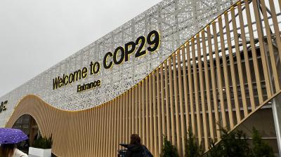 Entrance design for COP29 with a sign that reads "Welcome to COP29 Entrance," featuring wooden slats and a decorative perforated metal surface. People are walking nearby.