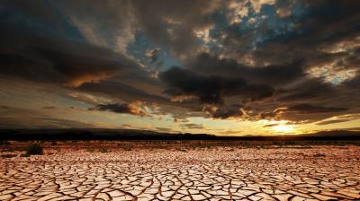 A cracked, dry desert landscape with dramatic clouds overhead and the sun setting on the horizon.