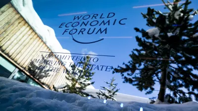 World Economic Forum sign on a glass panel, snow and trees reflected, with a snowy landscape and clear blue sky.