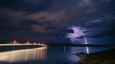 Lightning strikes over water near a long bridge with red lights under a stormy night sky.