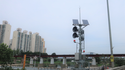 A tall pole with solar panels and loudspeakers stands by a river, near a railway bridge and high-rise buildings under a cloudy sky.