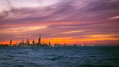 City skyline at sunset with colorful clouds above, a large body of water in the foreground, and a small boat on the water.