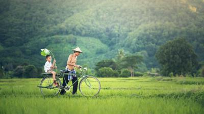 A shirtless man wearing a conical hat rides a bicycle through a lush green field, with a young boy holding a large leaf seated on the back.