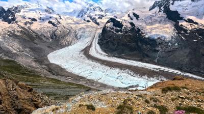 Mountainous landscape with a winding glacier flowing through rocky terrain. Snow-capped peaks are visible under a partly cloudy sky. Sparse vegetation is present in the foreground.