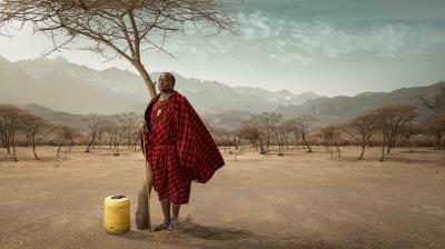 A person in traditional red attire leans against a tree in a dry landscape, with mountains in the background and a yellow container nearby.