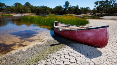 A red canoe sits on cracked, dry ground beside a shrinking body of water with sparse vegetation and trees in the background.