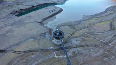 Aerial view of a partially dried reservoir with exposed land, visible tire tracks, and a circular water intake structure connected by a walkway.
