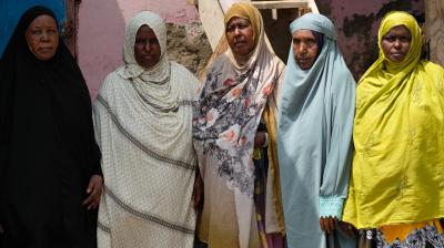 Five women in colorful hijabs stand side by side in front of a pink wall and wooden stairs, looking at the camera.