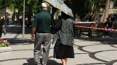 An older couple walks down a sunny, tree-lined sidewalk. The woman holds a large umbrella, and they are walking arm in arm away from the camera.