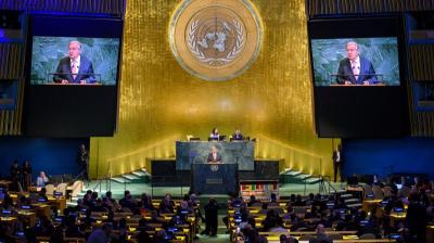 A speaker addresses delegates at the United Nations General Assembly hall, with two screens displaying his image and the UN emblem visible in the background.