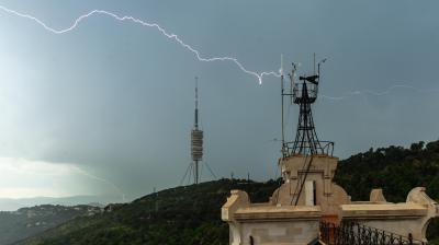A bolt of lightning strikes near a tall communications tower and an observation building on a hill, with dark clouds overhead and green landscape below.