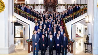 A large group of formally dressed people pose for a group photo on a grand staircase decorated with festive garlands in an ornate building lobby.