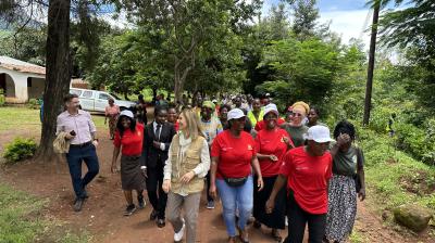 A group of people, including women in red shirts and white caps, walk together outside on a tree-lined path, with others and a white van visible in the background.