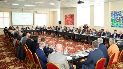 A large group of people sit around a U-shaped conference table in a meeting room, with a presentation slide displayed at the front.