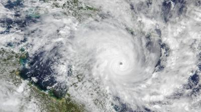 Satellite view of a large hurricane with a well-defined eye over the ocean, surrounded by thick clouds and spiral bands extending toward nearby landmasses.