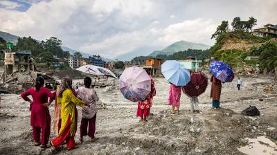 People holding umbrellas stand on a rocky, debris-filled landscape overlooking a town near mountains.