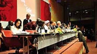 A group of people sits at a long table on a stage during a formal conference or panel discussion, with microphones and documents in front of them.