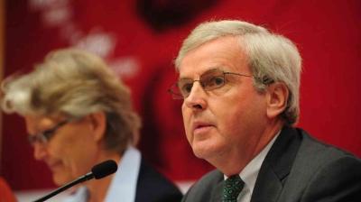 Two people sit at a table with microphones; the man in the foreground wears glasses and a suit, and the background is red and slightly blurred.