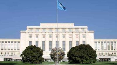 Large neoclassical building with a UN flag on top, a globe sculpture in front, and two trees on either side, set against a clear blue sky.