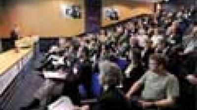 A large audience sits in rows facing a stage or panel in a conference or lecture hall.