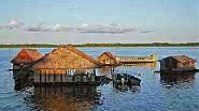 Thatched-roof huts and structures on stilts stand over a body of water, with a distant shoreline and blue sky in the background.