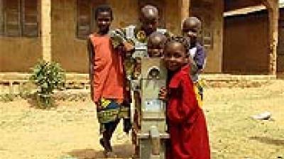 Five children stand around a manual water pump in an outdoor setting, with a building in the background.