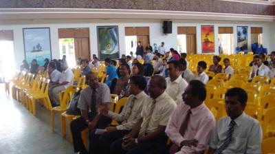 People seated on yellow chairs in a hall, with some attendees talking and others looking forward; wall art and windows are visible in the background.