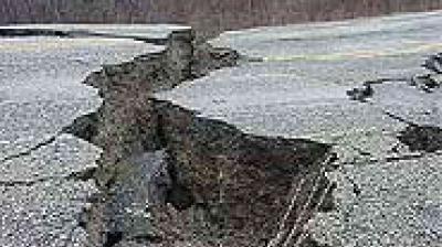 A large crack runs through the middle of a damaged asphalt road, showing signs of severe earthquake or ground movement.