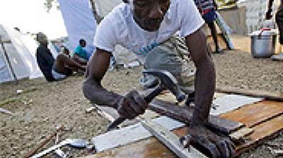 A man in a white shirt repairs wooden boards with a hammer outdoors, while others sit in the background on a gravel surface.