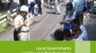 A group of people, some standing and some sitting on a road outdoors, next to a green banner titled "Local Governments and Disaster Risk Reduction.