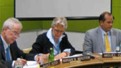 Three people in business attire sit at a table with nameplates, papers, and drinks in front of them, appearing to participate in a formal meeting.