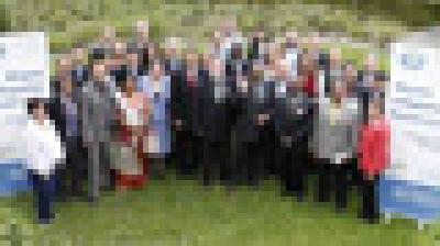 A large group of people poses outdoors on grass between two standing banners, with greenery in the background.
