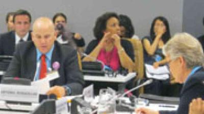 Several people sit at conference tables during a formal meeting, with microphones and documents in front of them.