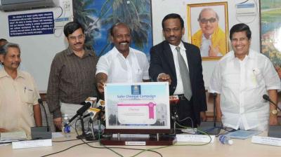 Five men stand around a table at a press event, unveiling a sign titled "Safer Chennai Campaign." Microphones and documents are on the table. A portrait hangs on the wall behind them.