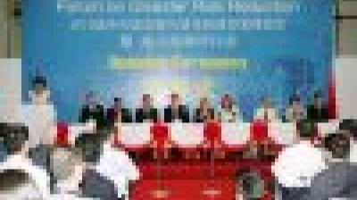 A group of people sit at a long table on a stage with a blue banner behind them at a formal event. Audience members are seated in front.