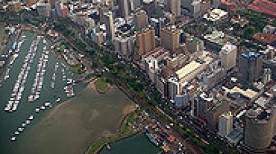 Aerial view of a coastal city with high-rise buildings, a marina filled with boats, and green spaces along the waterfront.
