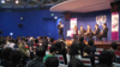 A large audience watches a panel of speakers seated on a stage in a blue-walled auditorium.