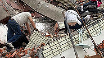 Two people search through debris and rubble of a collapsed building.