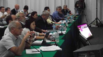 A group of people sit at tables with laptops, papers, and water bottles, attending a conference or meeting; a presentation screen is visible in the foreground.