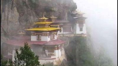A monastery with golden roofs is perched on a foggy mountainside, partially obscured by mist, with rocky cliffs and greenery visible.