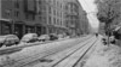 A city street in winter with snow covering parked cars, tram tracks, and sidewalks, lined with multi-story buildings and bare trees.