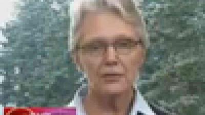 A pixelated image of a person with short light hair and glasses, standing outdoors with blurred trees in the background.