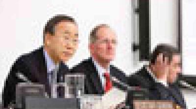 Three men in suits sit at a conference table with microphones and nameplates, appearing to participate in a formal meeting or panel discussion.